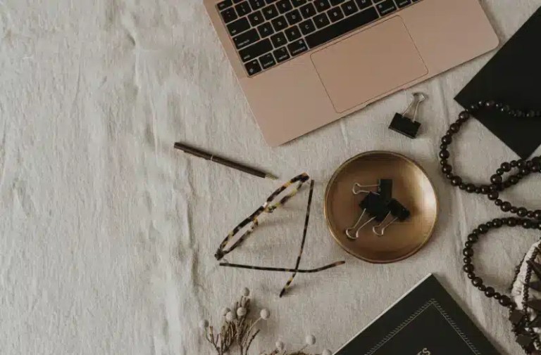 A flat lay of a workspace with a rose gold laptop, eyeglasses, a pen, binder clips in a brass dish, and a closed notebook on a neutral fabric surface, styled with beads and dried flowers for a calm digital detox vibe.