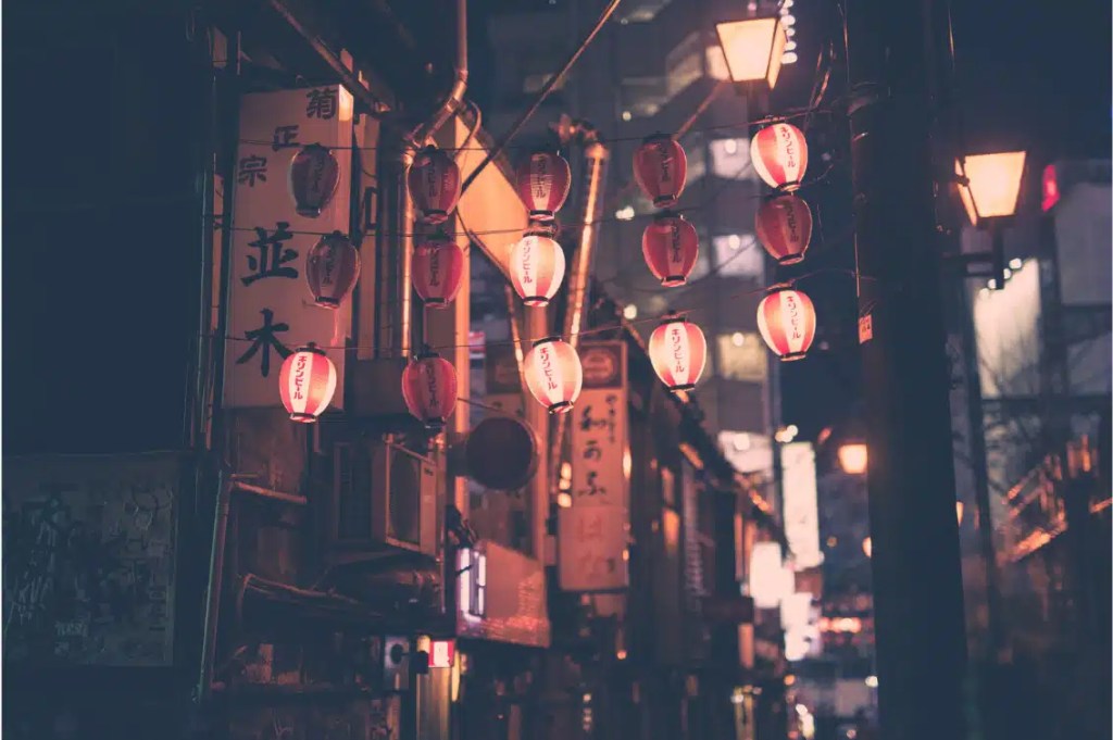 A narrow alleyway in Japan illuminated by traditional red and white paper lanterns at night, with warm lighting and urban buildings in the background.