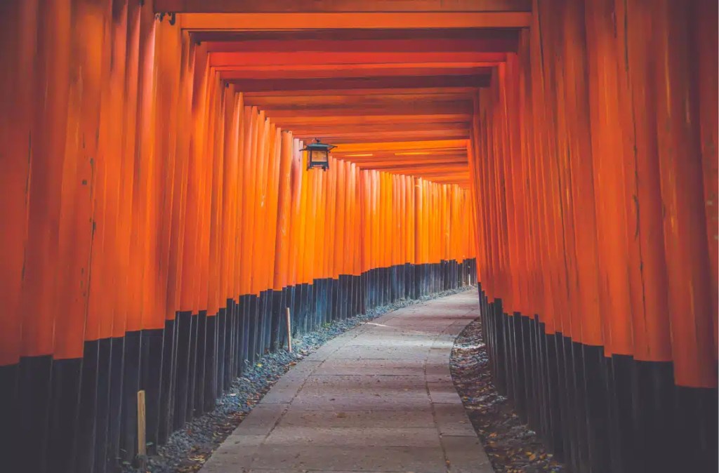 A winding pathway through the iconic vermillion torii gates of Fushimi Inari Shrine in Kyoto, Japan, creating a tunnel-like effect with vibrant orange and black hues.