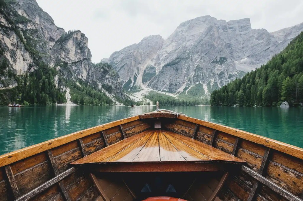 The view of misty grey mountains from the point of view of a wooden row boat on a calm blue lake. The sky is white, suggesting a cold day, while the lake and mountains are trimmed with green pines and trails of white snow.