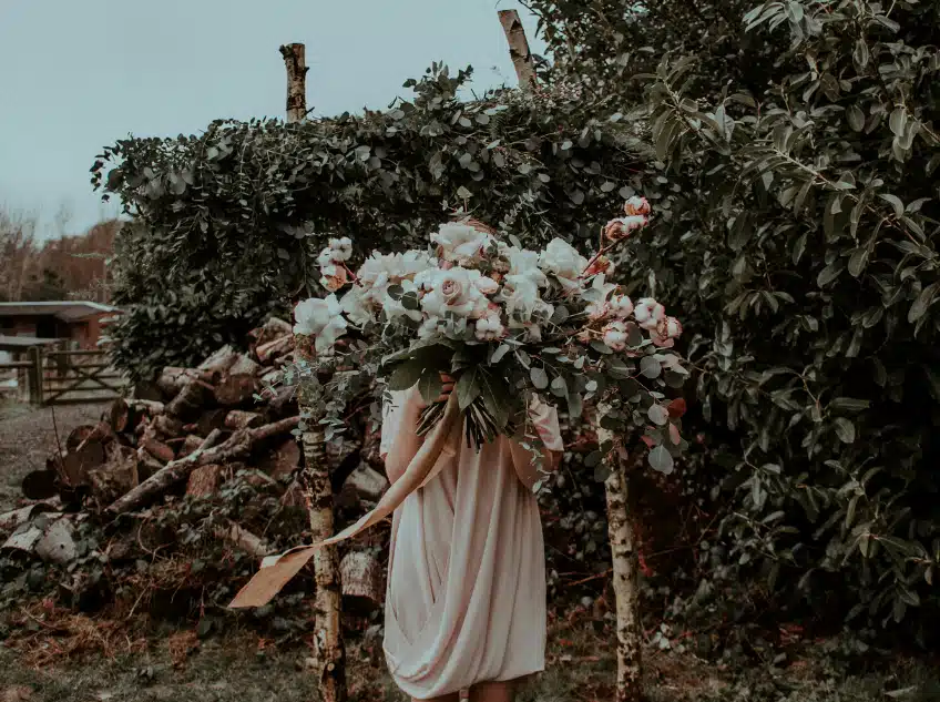 A woman in a soft, flowy dress holds an oversized bouquet of roses, eucalyptus, and greenery as she stands in a rustic outdoor setting with stacked firewood and lush foliage in the background.