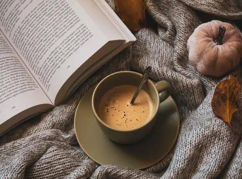 A cozy scene with an open book, a cup of coffee on a saucer with a spoon, a knitted gray blanket, dried autumn leaves, and a small fabric pumpkin, symbolizing relaxation and digital detox.