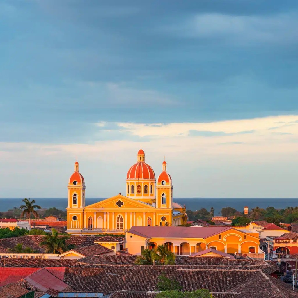 A view of the bright yellow Granada Cathedral with its striking red domes, set against a backdrop of rooftops, trees, and Lake Nicaragua in the distance.