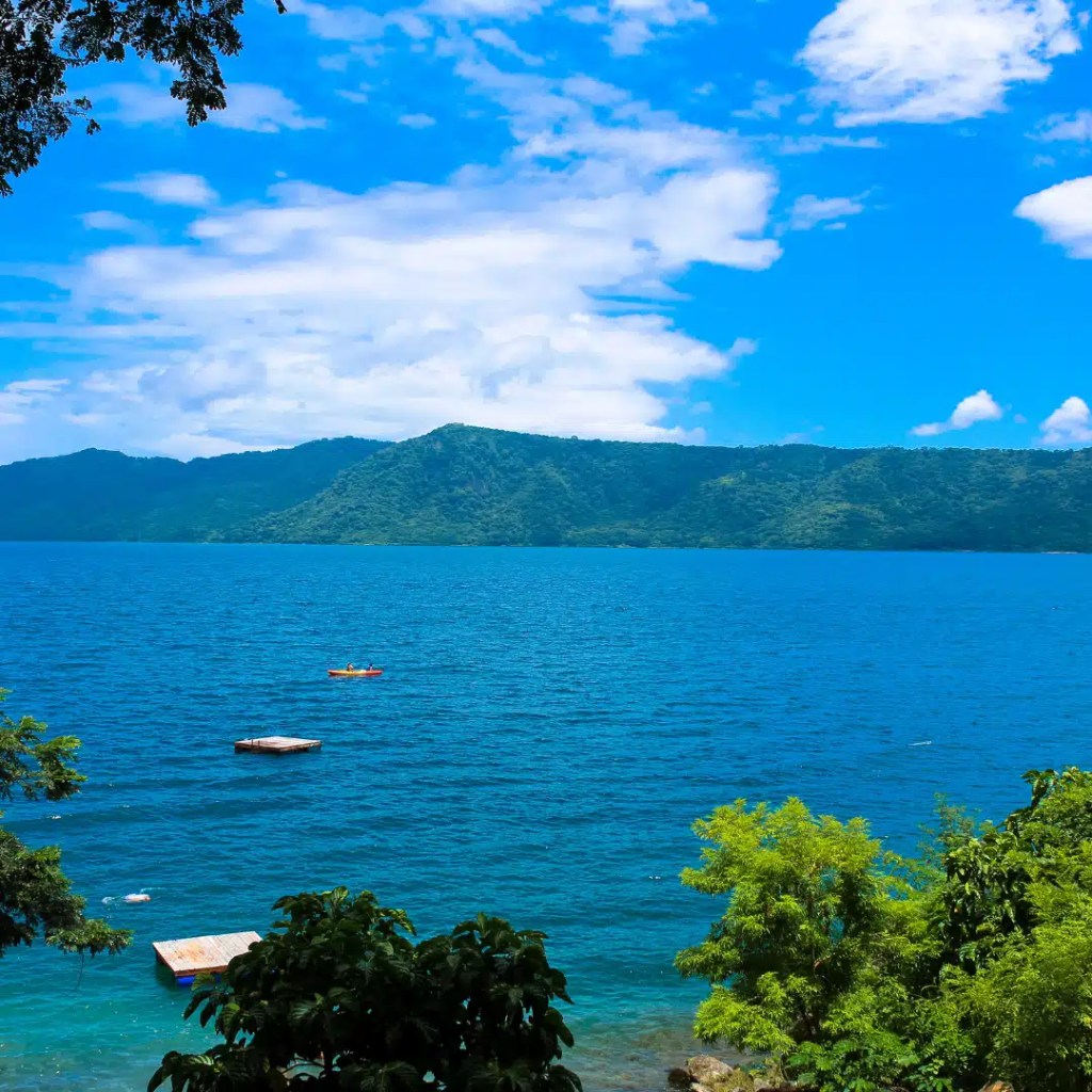 A panoramic view of Laguna de Apoyo, a crater lake near Granada surrounded by heavy rainforests. Lush green trees give way to the calm blue waters of the lake with vegetation covering the mountainous terrain in the background. A few small boats can be seen on the water.