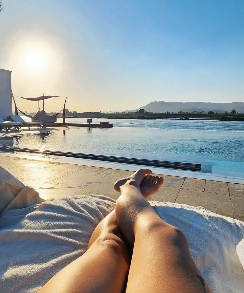You see me relaxing by a serene infinity pool at sunrise, with legs stretched out on a lounger overlooking the Nile River and distant hills in Luxor, Egypt. The soft sunlight casts a golden glow across the water, creating a peaceful and tranquil atmosphere.