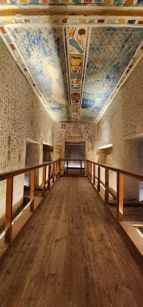 An interior view of an ancient Egyptian tomb in Luxor, Egypt, featuring a wooden walkway leading through a narrow, decorated corridor. The walls are covered with hieroglyphs and colorful artwork, while the ceiling showcases a stunning blue and gold celestial design with vibrant depictions of deities and symbols.