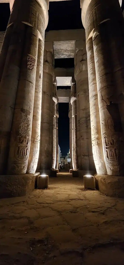 A night-time photo of the Luxor Temple in Egypt. The image shows a long, covered walkway lined with tall, intricately carved columns. The columns are lit by spotlights, casting a warm glow on the ancient stone. The walkway leads towards a dimly lit area in the distance.