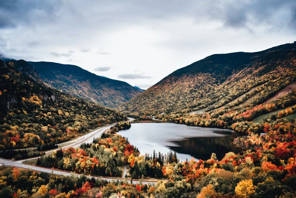 A New England lake surrounded by mountains covered in autumn trees, with a road curving along the shoreline