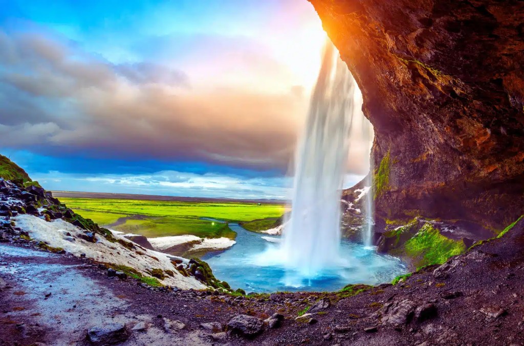 Walk behind Seljalandsfoss waterfall in Iceland, on a partly cloudy day with green fields in the distance.