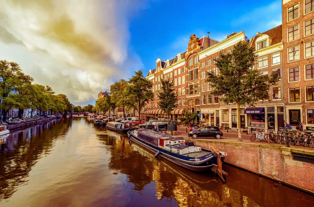 View of a picturesque Amsterdam canal with traditional houses, houseboats, trees, and bicycles on a sunny day.