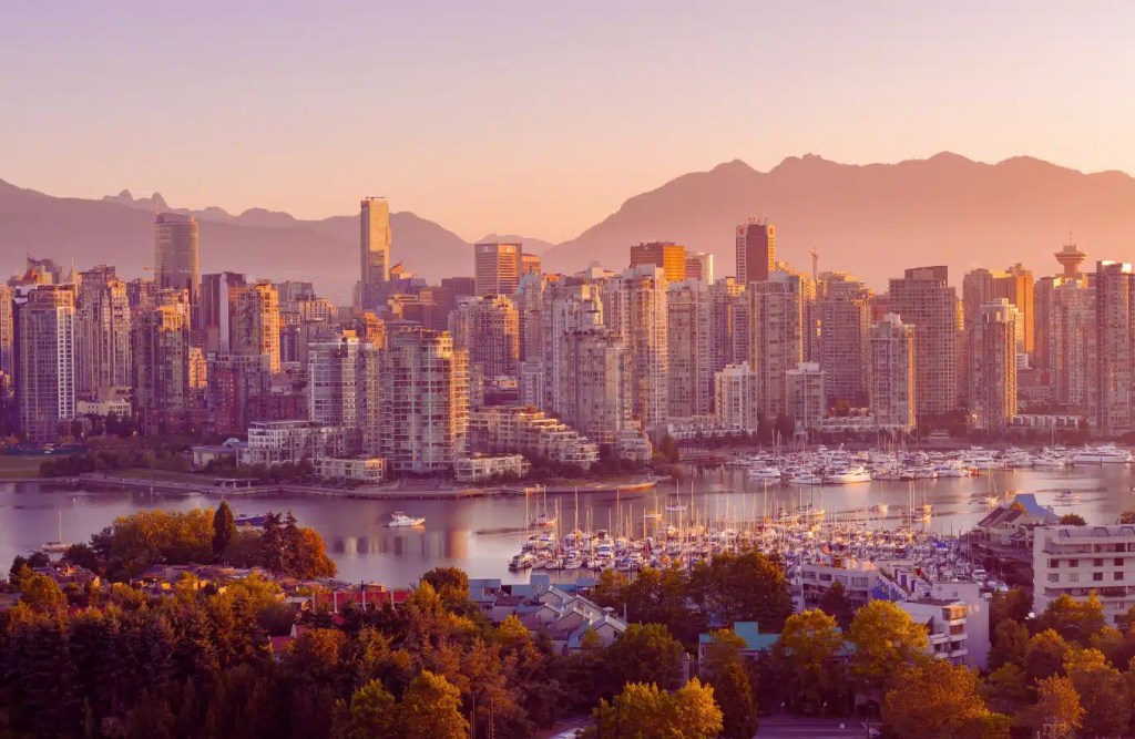 Vancouver skyline at sunset, with sailboats moored in the harbor and mountains in the background.