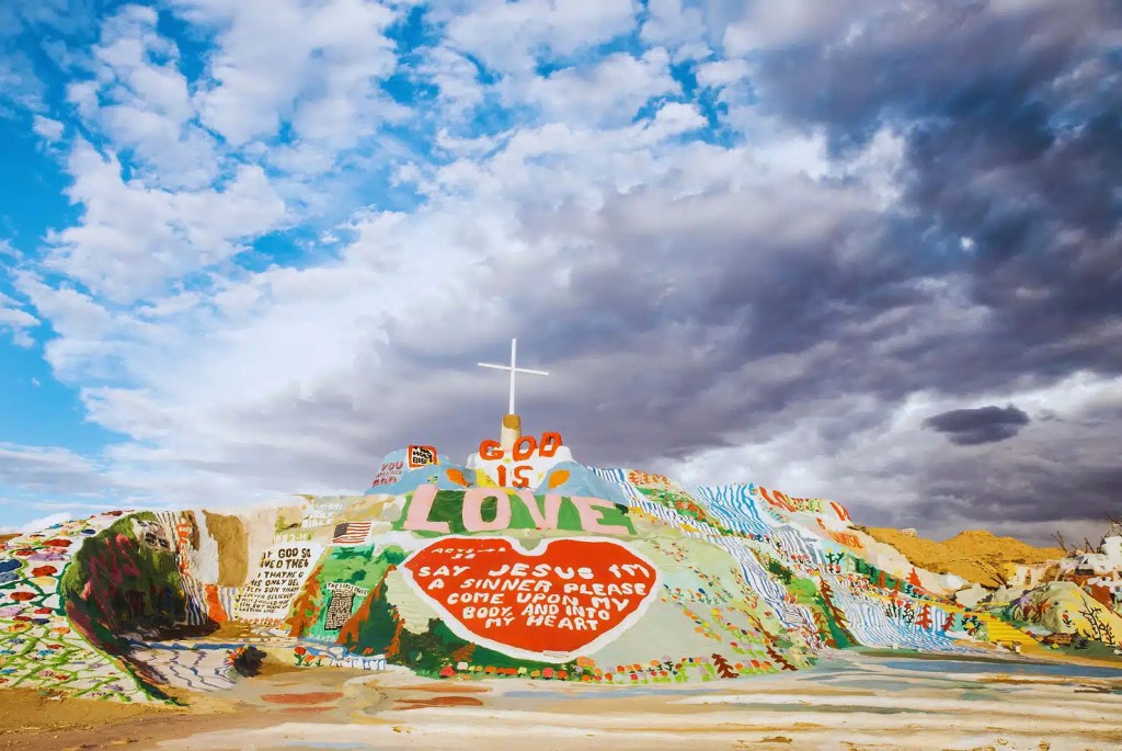 Salvation Mountain in California, a colorful hillside covered in murals and religious messages with a white cross at the top, set against a dramatic cloudy sky.