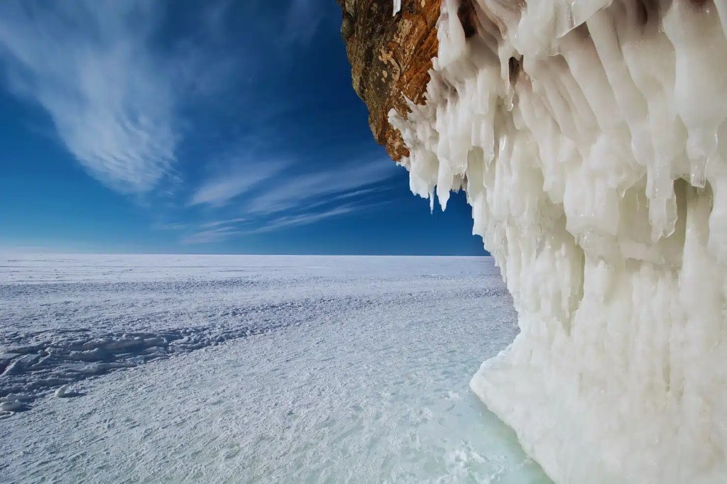 An interesting view of Apostle Islands National Lakeshore in winter. A sandstone sea cave is completely frozen over, with icicles dripping from the ceiling and the cave entrance blocked by ice. The vast expanse of Lake Superior is frozen and covered in snow, stretching out to the horizon under a bright blue sky.