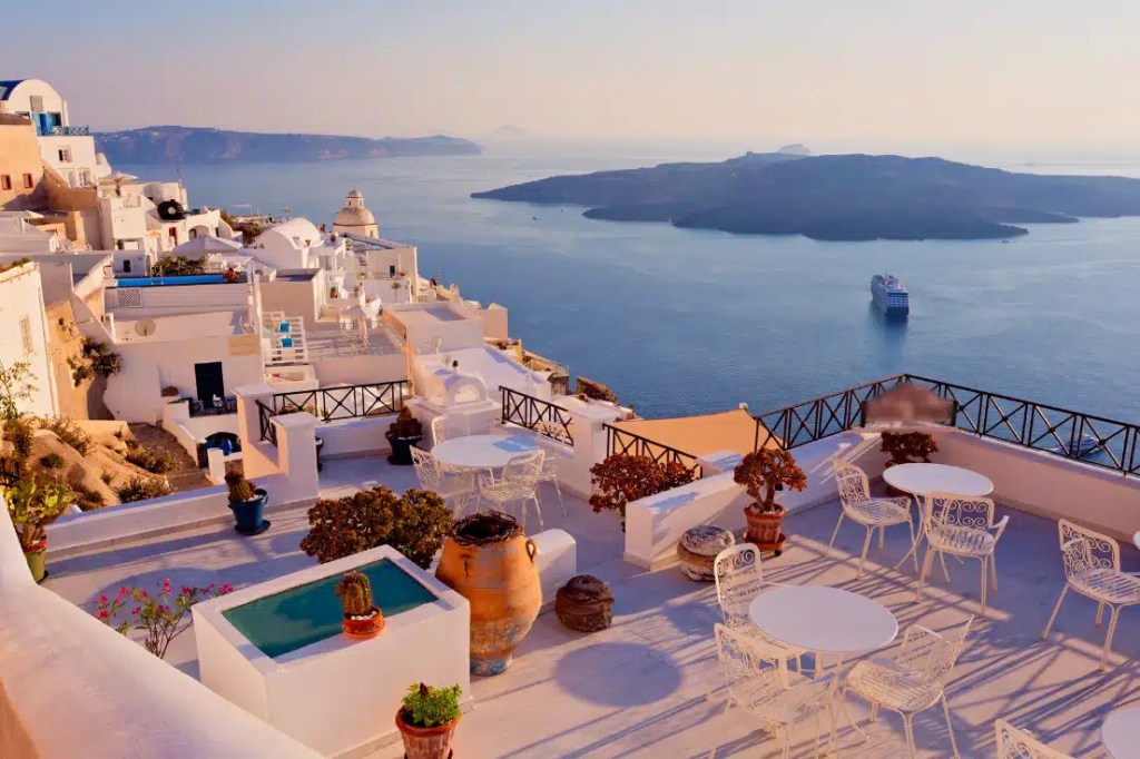 The image showcases a beautiful terrace in Santorini, Greece, with whitewashed buildings and an iconic view of the calm blue Aegean Sea. The terrace features white metal tables and chairs, surrounded by potted plants, and overlooks the caldera, with a large island and a cruise ship in the distance. The sky is soft with pink and purple hues, suggesting early morning or sunset.