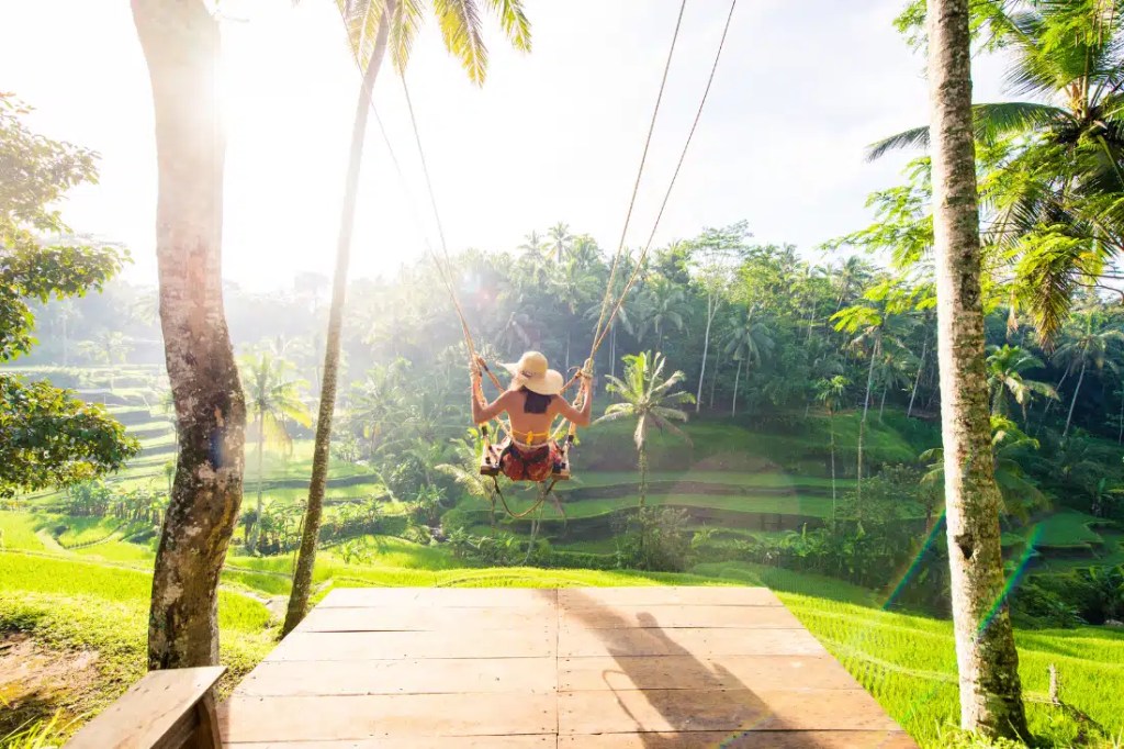 The image shows a person on a large swing, suspended between 2 palm trees, overlooking the lush green rice terraces of Bali, Indonesia. The person, wearing a hat and light summer clothing, swings high into the air, with the sun shining brightly through the trees, creating a dreamy atmosphere.