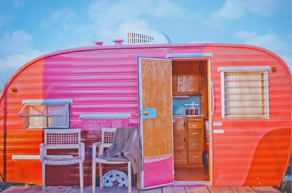 A vintage, candy-colored pink trailer parked in Marfa, Texas, with its door open revealing a cozy interior. Two chairs and a blanket are on the porch, inviting relaxation in the desert landscape.
