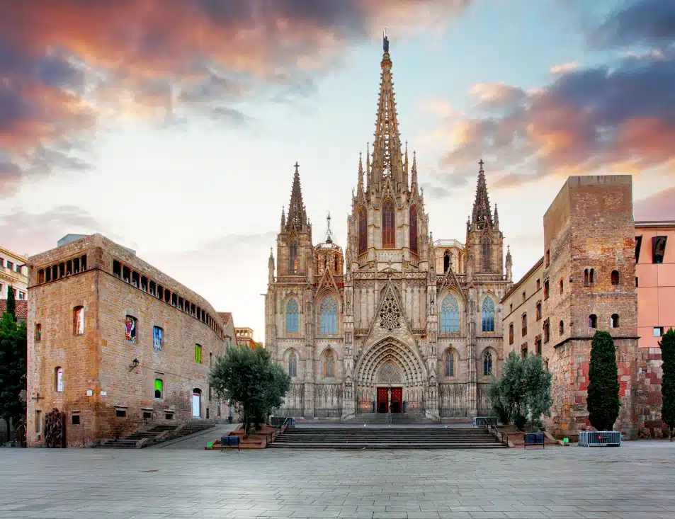 An image of the Barcelona Cathedral, showcasing its stunning Gothic architecture. The façade features intricate details, spires, and a large central entrance, with the sky displaying warm tones of sunset in the background. The cathedral is situated in a spacious, empty square with surrounding historical buildings.