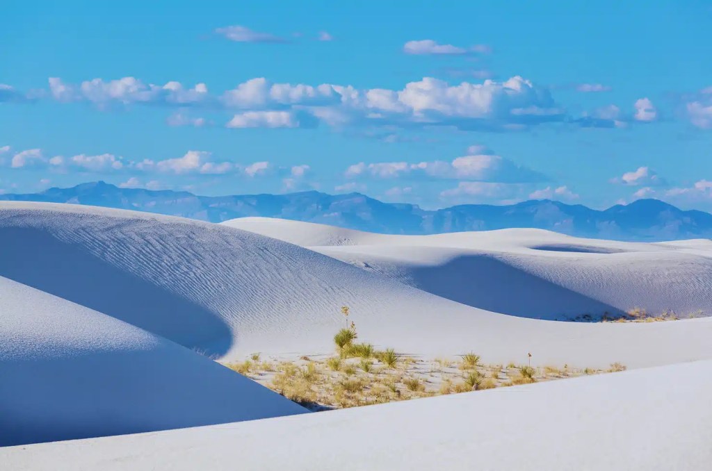 A vast expanse of white sand dunes stretches across the horizon under a bright blue sky at White Sands National Park. Wispy clouds drift overhead, casting shadows on the dunes.