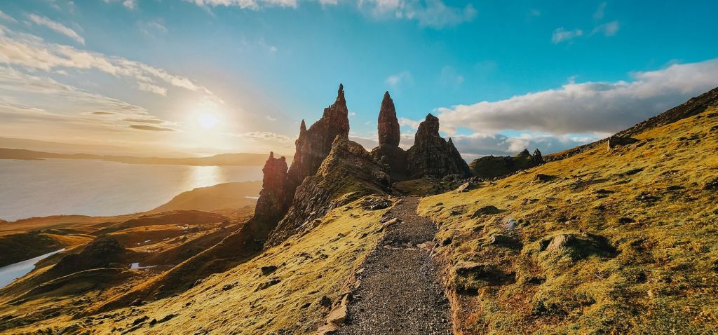 Pathway leading to the Old Man of Storr on the Isle of Skye, Scotland, with jagged rock formations and vibrant green hills illuminated by the golden glow of the sunrise.