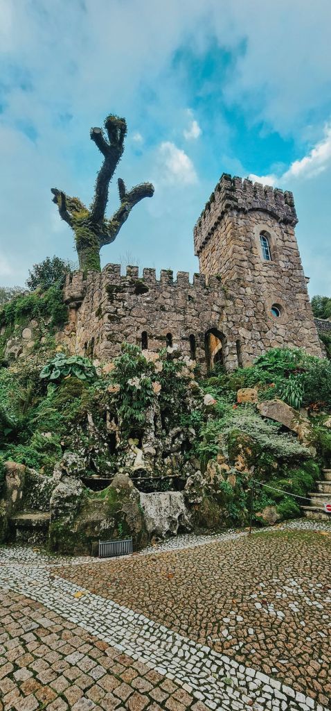 A stone medieval-style tower surrounded by lush greenery and cobblestone paths in Sintra, Portugal, set against a bright, partly cloudy sky.