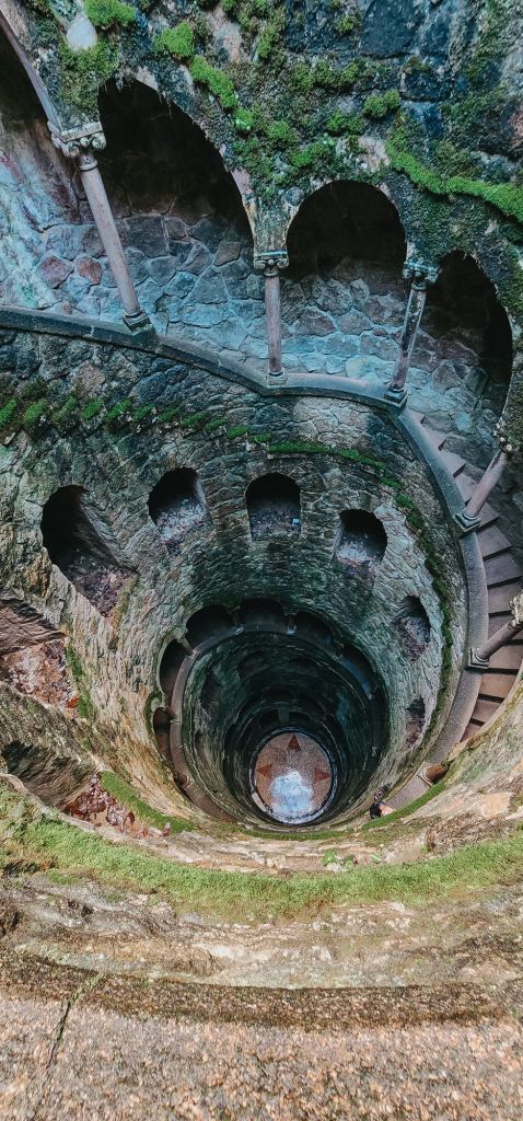 The spiraling stone staircase of the Initiation Well at Quinta da Regaleira in Sintra, Portugal, descending into a shadowy underground structure with moss-covered walls and arched openings.