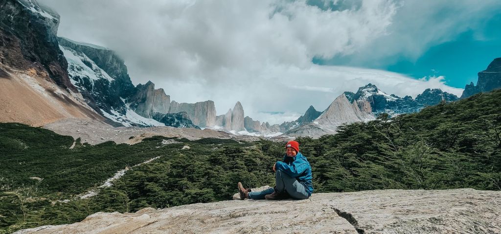 Here I am as I hike the W Trek in Patagonia, sitting on a rocky outcrop with a scenic view of the rugged mountains and thick forest in Torres del Paine National Park, under a partly cloudy sky.