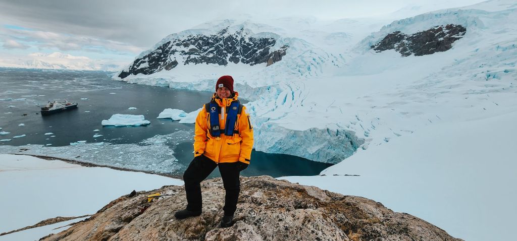 This is an image of me standing on a rocky outcrop in Antarctica, wearing a bright yellow expedition jacket, blue life vest, and a red hat. Behind me, you can see a stunning landscape of glaciers, icebergs, and snow-covered mountains, with a Quark Expeditions ship visible in the icy waters below.